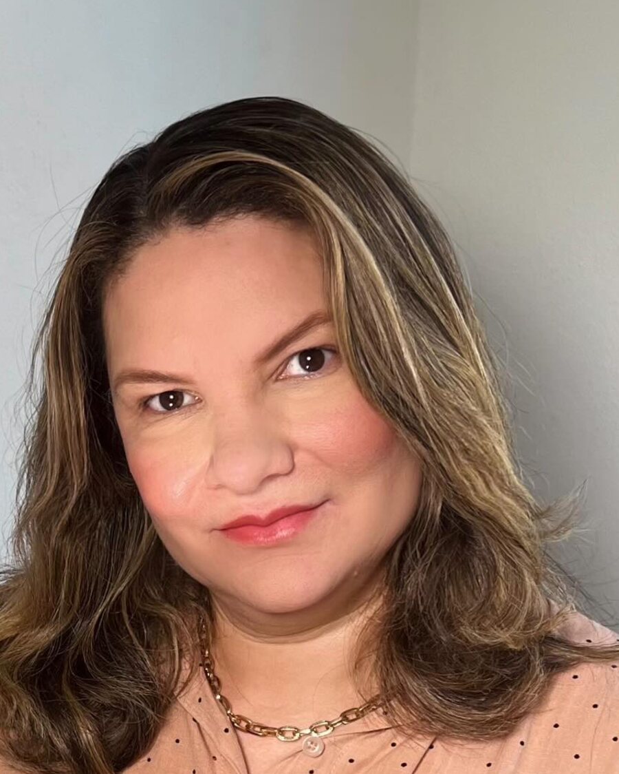 Dra. Laucimar Rodrigues na clínica, de blusa rosé, olhando para a câmera.
